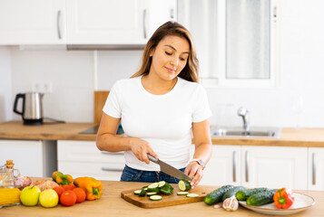 Obraz na płótnie Canvas Positive young latin american girl standing in cozy kitchen interior at home, slicing fresh vegetables for salad