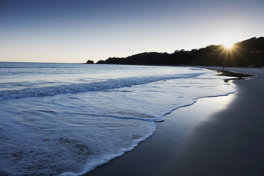 Surf on shoreline of beach with sun shining over silhouetted hillside at Byron Bay in New South Wales, Australia
