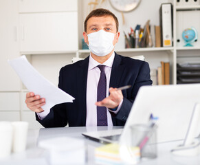 Focused male in disposable face mask working in business office using laptop, new normal due to...