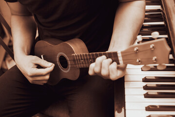 A young man holds a small ukulele guitar in his strong hands and learns to play it, next to the piano, a guy has an online ukulele lesson