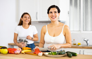 Cheerful young Hispanic woman with female roommate cooking together in home kitchen, preparing vegetable dish for dinner