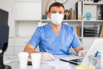 Fototapeta na wymiar Focused man doctor wearing medical facial mask and gloves working in medical office using laptop computer
