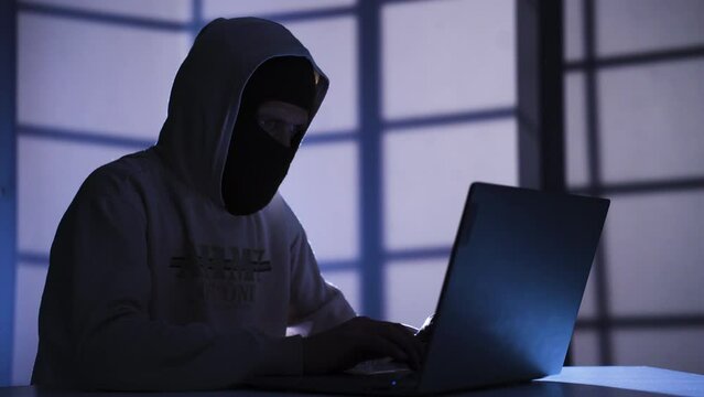 Male in ski mask hacking in a dark room.
Mature male hacker commits cyber crime in dark room, hacking online sites.
