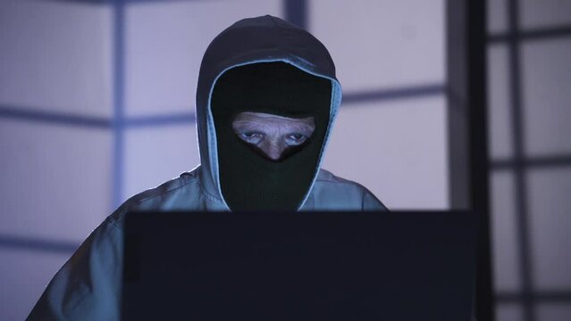 Masked hacker working from laptop to find security password.
Masked dangerous hacker cracking password from laptop in dark office, hacking online sites. Cyber attack concept.
