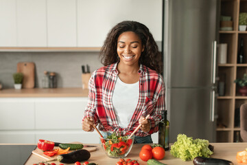 Satisfied glad millennial black female preparing salad at table with organic vegetables, has fun at...