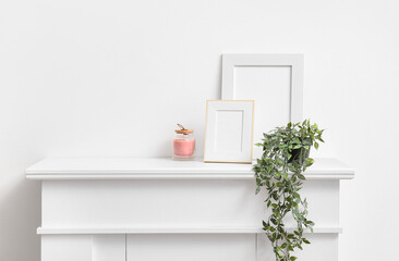 Blank photo frames, candle and houseplant on mantelpiece near white wall