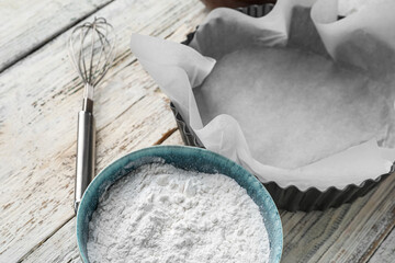 Baking dish with paper, bowl of flour and whisk on white wooden background, closeup
