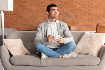 Young man with cup of tea sitting on grey sofa at home