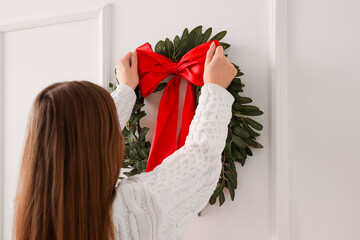 Young woman hanging Christmas wreath on light wall in living room, closeup