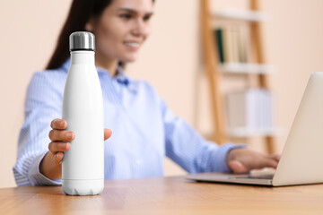Young woman taking thermo bottle at workplace indoors, focus on hand