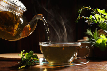 Pouring aromatic herbal tea into cup, mint and rosemary on wooden table