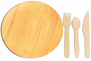A set of eco-friendly cutlery: wooden fork, spoon and knife next to a biodegradable wooden plate on a white background