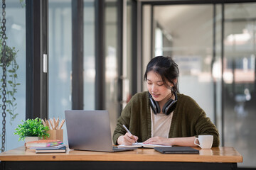 Asian businesswoman making notes looking at a laptop and wearing headphones.