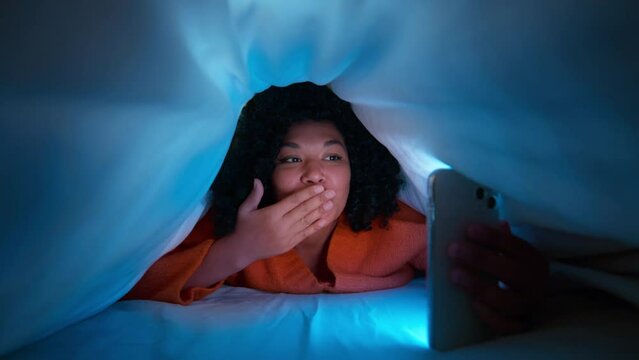 Happy African American female under blanket. Woman of color with afro hair taking pictures for social media or talking with friend using smartphone video conference calling laying on bed at night 4K