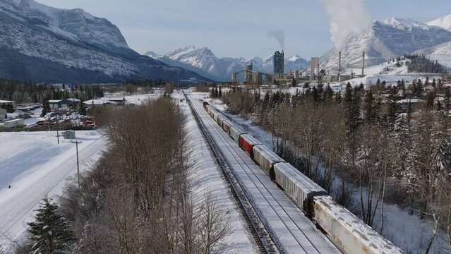 Aerial View Of Parked Freight Train With Snow Covered Landscape Near Lafarge Exshaw Cement Plant And Snow Capped Mountains In The Distance. Dolly Forward