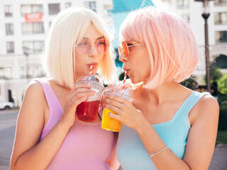 Two young beautiful smiling hipster female in trendy summer clothes. Sexy carefree women posing in the street in wigs. Positive funny models drinking fresh cocktail smoothie drink in plastic cup