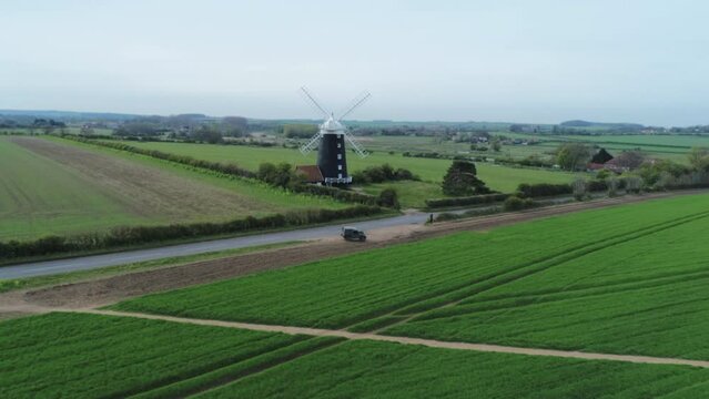 Rising Aerial Drone Shot Of Rural Old 19th Century Grade II Listed Windmill and Green Fields with Old Landrover Defender Parked on Side of Road Burnham Overy Staithe North Norfolk UK