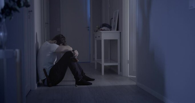 Man sitting on the floor of his home depressed, squatting with a bottle of super alcohol in his hand. Static view