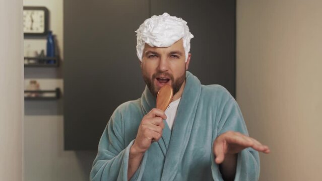 A man with a foam hairdo sings into the brush wearing a bathrobe