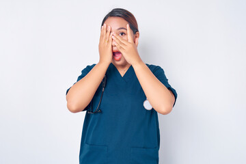 Scared and shocked young Asian woman nurse wearing blue uniform with a stethoscope witness something embarrassing or scary, cover her eyes but peek through finger isolated on white background