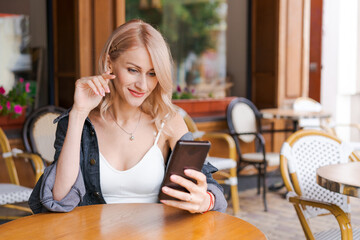 Young cute woman using phone sits in cafe at table with smartphone, answering texts, phone calls, letters, posting photos on Instagram, outdoor portrait, close-up caucasian girl