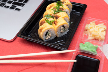 Delicious Japanese rolls with salmon, avocado, cucumber, cheese in takeout packaging with wood sticks, soy sauce and laptop on office table. Selective focus. Asian delivery food concept