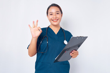 Smiling young Asian woman nurse wearing blue uniform with a stethoscope holding clipboard and showing okay sign isolated on white background. Healthcare medicine concept