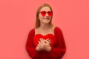 Excited dreaming happy young woman holding red toy heart close to chest, wearing red dress with...