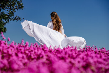 a beautiful woman in a white flowing long dress stay near a beautiful field with pink flowers rear...
