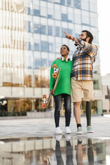 Obraz premium funny couple with skateboard, in the city standing pointing, african american woman, hispanic man with amputated leg and a prosthesis.