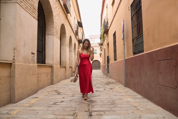 Fototapeta na wymiar Young, attractive, blonde woman, wearing an elegant red party dress and holding golden high heels in her hand, walking barefoot down a city alley. Concept beauty, fashion, elegance, luxury.