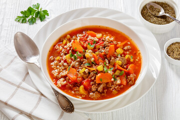 Easy hot and hearty Hamburger Soup with barley and vegetables in white bowl on white wood table, horizontal view from above
