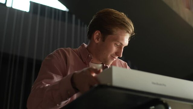 Ufa, Russia - 10 October 2022. Close-up low-angle view of satisfied smiling young man unboxing cardboard box Apple MacBook Pro 16 inch sitting on sofa at table in sunny living room. Online shopping