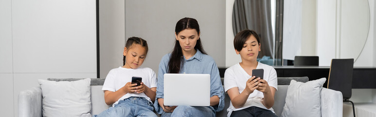 Mother working on laptop near asian kids with smartphones at home, banner
