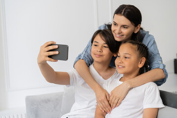 Smiling mother hugging asian kids while son taking selfie at home