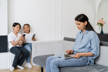 Mother using laptop near blurred asian children with cellphones at home