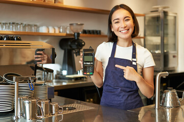 Smiling asian barista girl, wears apron, shows credit card machine for processing payment, suggest...