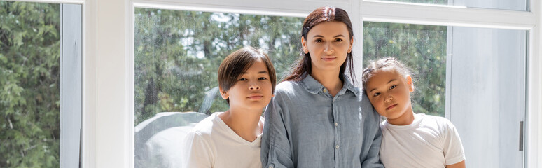 Obraz premium Mother and asian children looking at camera near window at home, banner