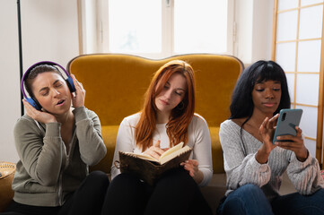Portrait of three multiracial female friends sitting in living room. Each woman is doing something...