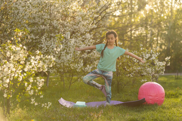 Little girl child doing yoga exercise stretching on the grass in sunny summer day. A flexible child, doing gymnastics exercises. Sports, learning, fitness, stretching, yoga, active lifestyle concepts.