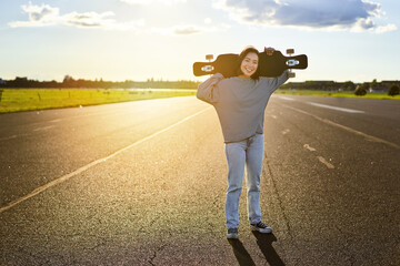 Young asian woman standing with longboard on sunny road, skating in skate park on her cruiser