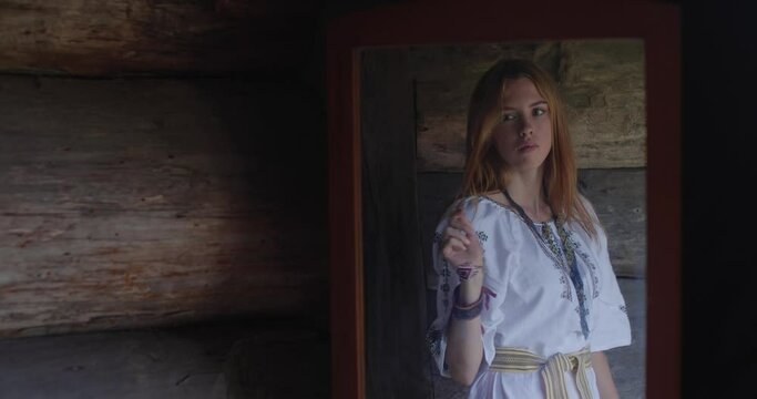 Attractive young blonde woman in an embroidered shirt looks into a mirror in an old wooden house, smiles and reaches out to touch her reflection. Divination, mysticism, magic concept.