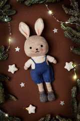 Cute knitted toy rabbit in a sweater and shorts on a Christmas background with fir branches and lights. A toy made by hand, a New Year's gift with your own hands.