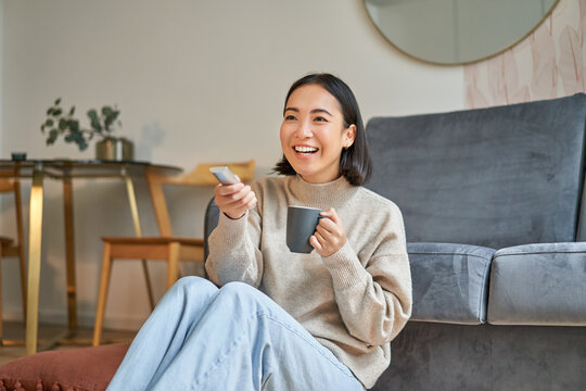 Portrait Of Beautiful Asian Girl Sitting At Her Home And Watching Tv, Holding Remote, Smiling And Laughing, Feeling Comfort And Warmth At Her Apartment