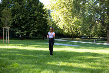 Fototapeta premium Woman walks in the park and reads a message on the smart phone. Portrait woman with brown hair, cup of coffee and mobile phone. Outdoor, public park, relaxed position and summer. 