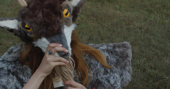 Close up portrait  of blonde girl in a pagan animal-like  fur costume with bone bell on her neck made of animal skins standing  against the grass backdrop putting on a goat mask with horns.