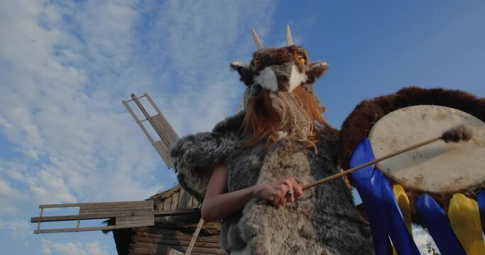 A blonde girl in a pagan fur costume made of animal skins and a goat mask with horns knocks on a leather tambourine against the background of an old wooden windmill and a rural landscape.