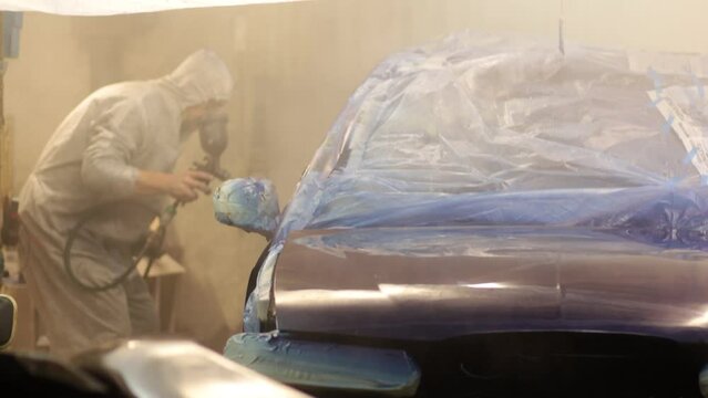 man in uniform paints a car blue with a spray gun in a garage. Coating the body of the car with varnish.