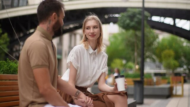 Happy pretty young man and woman having carefree talk sitting on bench holding hands with takeaway coffee looking at each other. Loving couple drinking coffee sitting on bench during summer day.