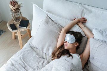 Sleeping young woman laying on bed, shoot from top. Calm caucasian girl having nap wearing sleeping mask resting at home at weekend. Fit Italian female relaxing at hotel on vacations. Rest and leisure